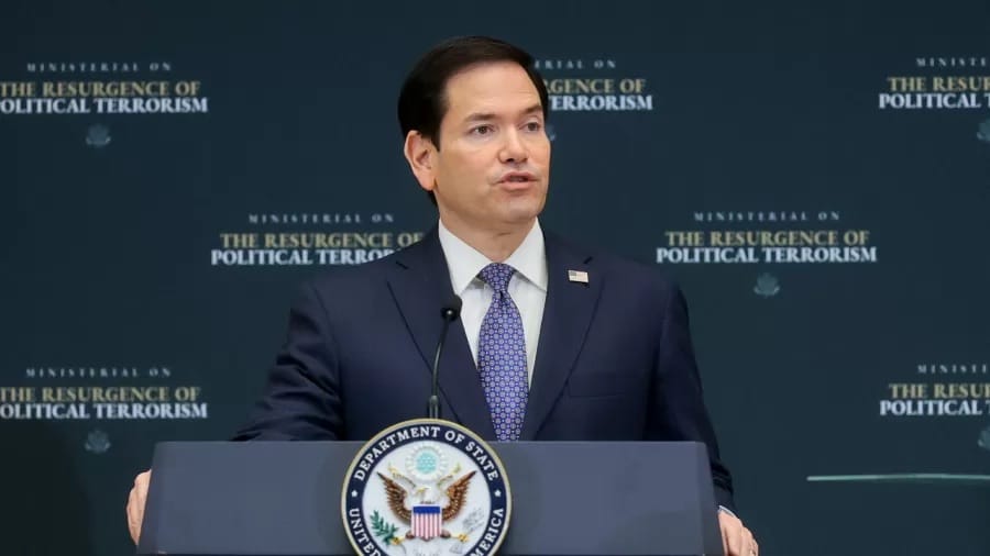 Marco Rubio, de terno azul-marinho e gravata azul, fala em um púlpito com o selo do Departamento de Estado diante de um painel escuro