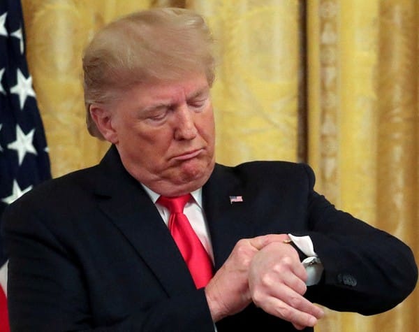 Presidente Donald Trump, de terno escuro e gravata vermelha, olha para o relógio de pulso com expressão fechada, diante da bandeira americana
