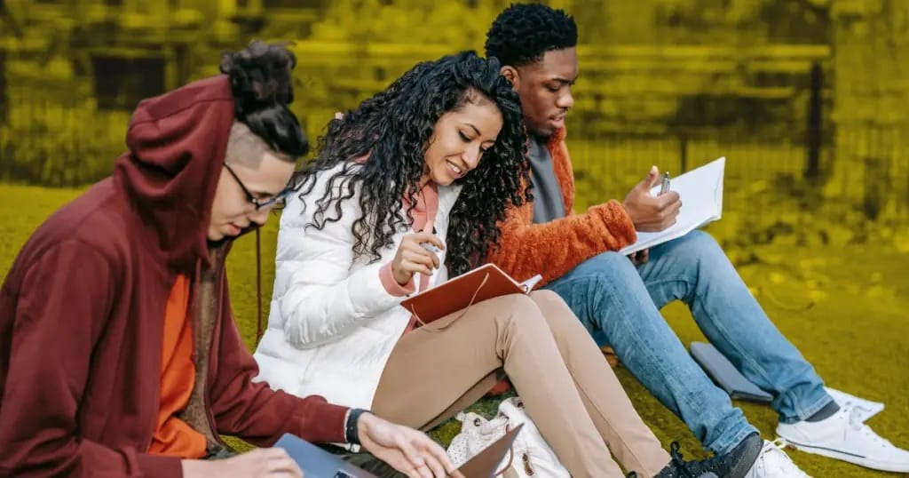 Três jovens sentados ao ar livre, com cadernos e canetas nas mãos, estudando juntos sobre um gramado