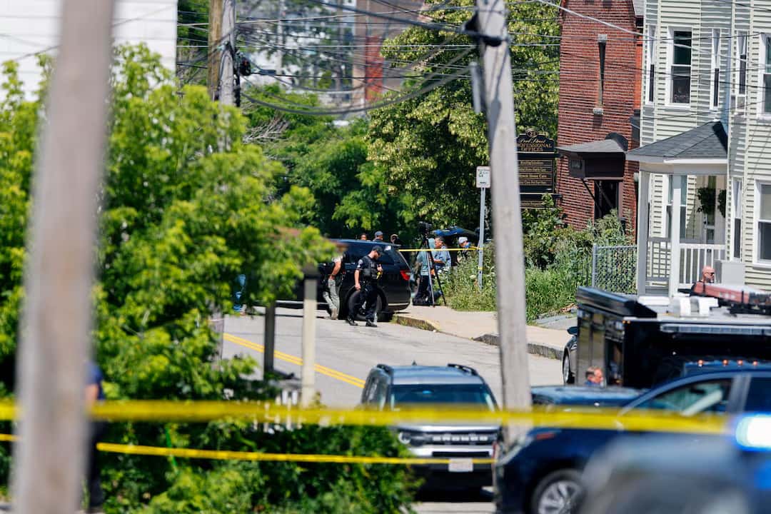 Rua residencial isolada por fita amarela de perícia, com agentes e viaturas ao fundo, no local onde um homem foi morto a tiros pelo ICE em Biddeford, no Maine