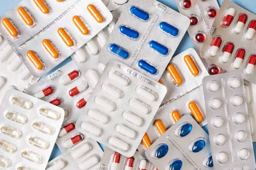 Várias cartelas de comprimidos e cápsulas coloridas, laranjas, azuis, brancas e vermelhas, espalhadas sobre uma superfície azul-clara