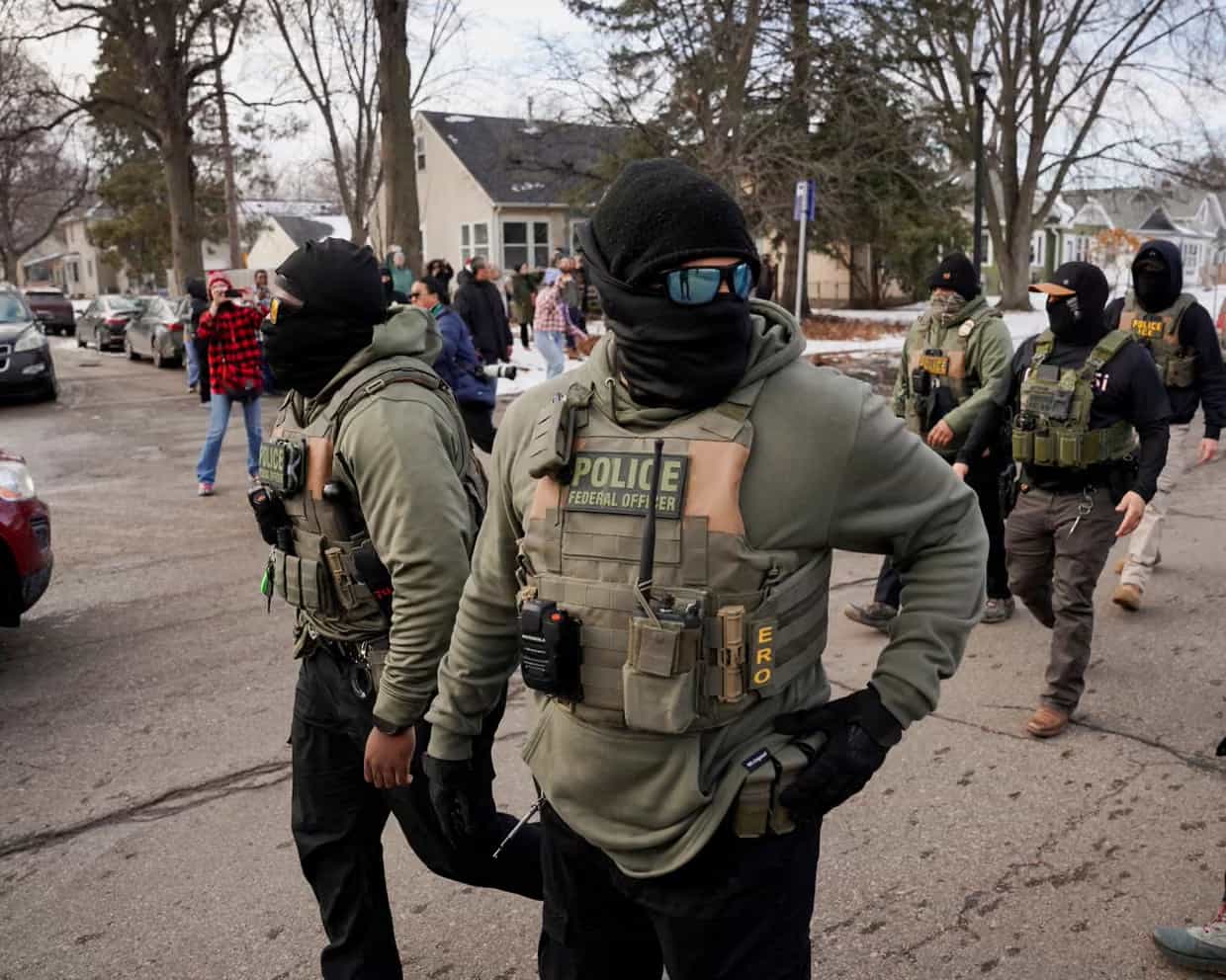 Agentes federais encapuzados com coletes onde se lê POLICE FEDERAL OFFICER e ERO caminham por uma rua residencial enquanto moradores observam e filmam ao fundo