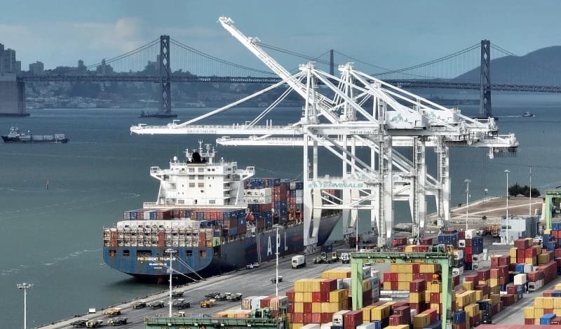Navio porta-contêineres atracado em terminal portuário com guindastes brancos e pilhas de contêineres coloridos, e a ponte da baía de São Francisco ao fundo