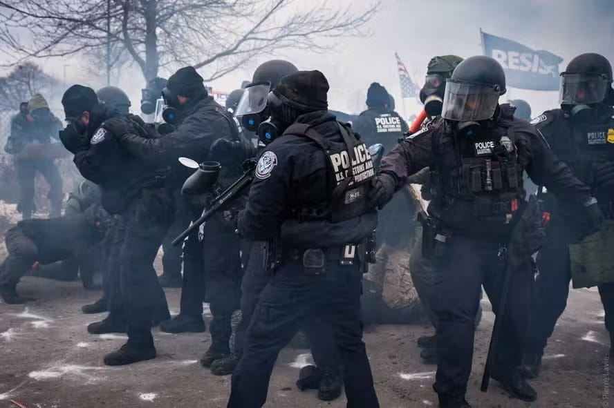 Fileira de agentes federais com máscaras de gás, capacetes e escudos em meio à fumaça de gás lacrimogêneo, com uma bandeira onde se lê RESIST ao fundo