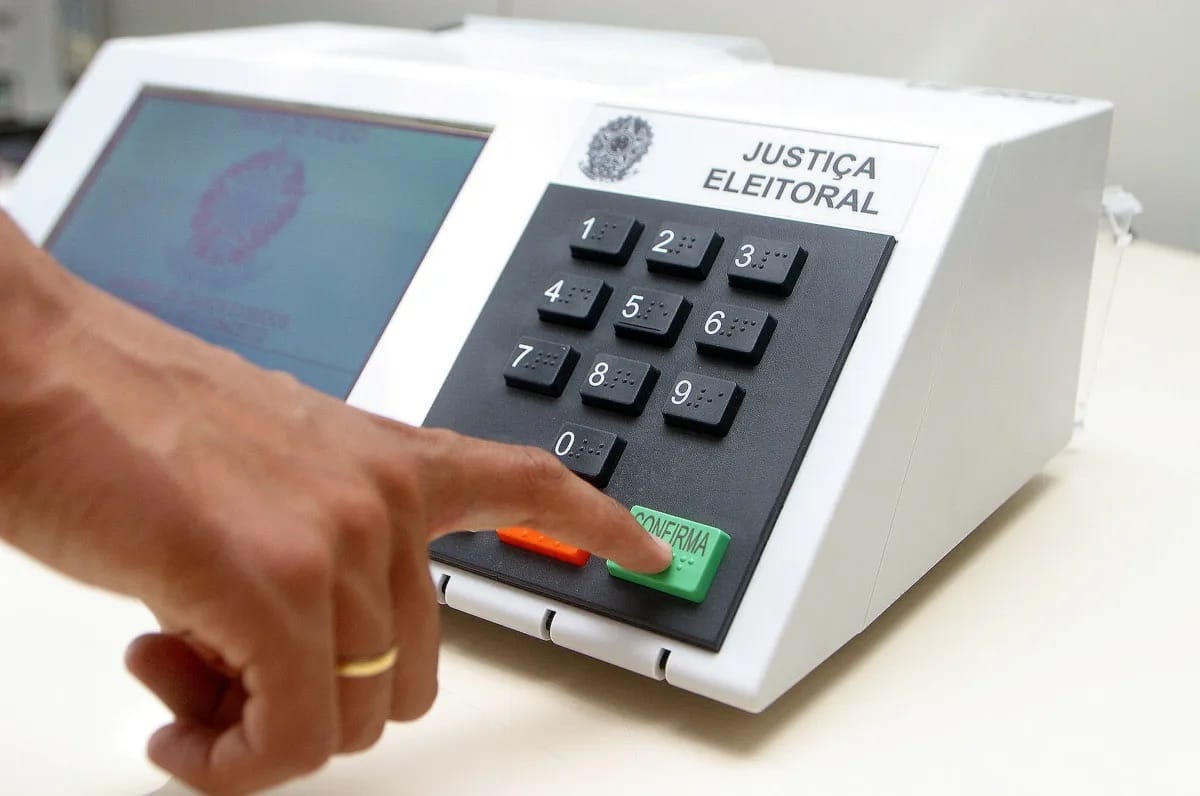 Mão pressiona a tecla verde de confirmação de uma urna eletrônica branca com o brasão da Justiça Eleitoral do Brasil