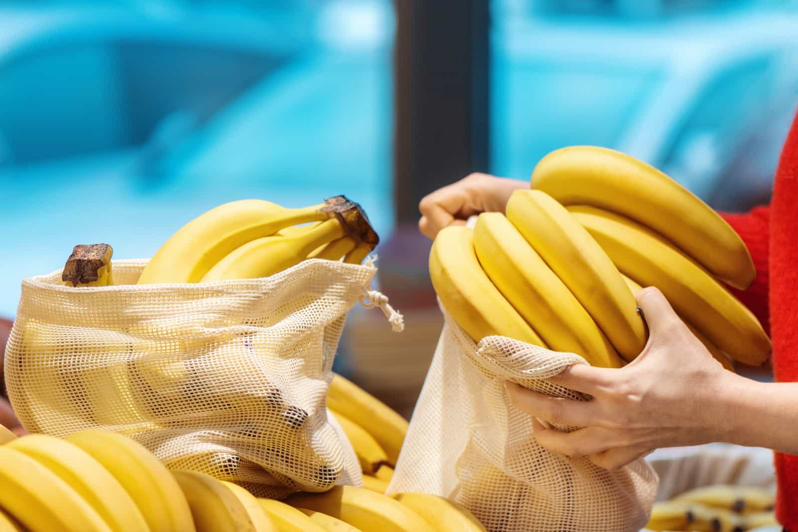 Mãos pegam cachos de bananas amarelas em sacos de rede, em uma banca de supermercado