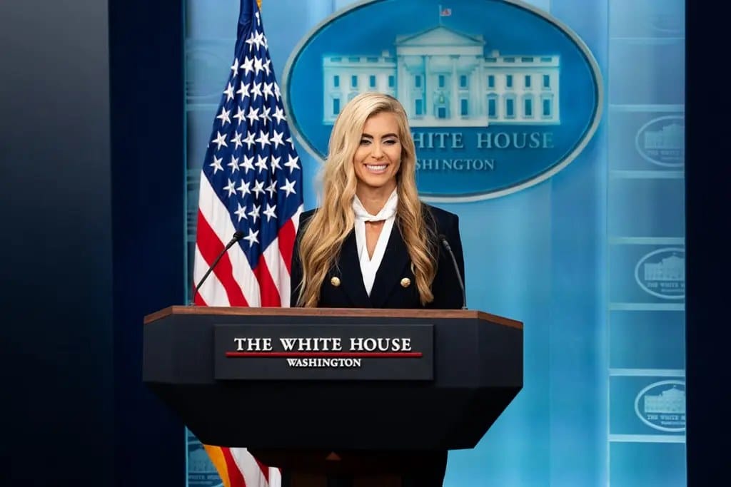 Mulher loira, de blazer escuro, sorri atrás de um púlpito com a inscrição The White House, diante de uma bandeira dos EUA
