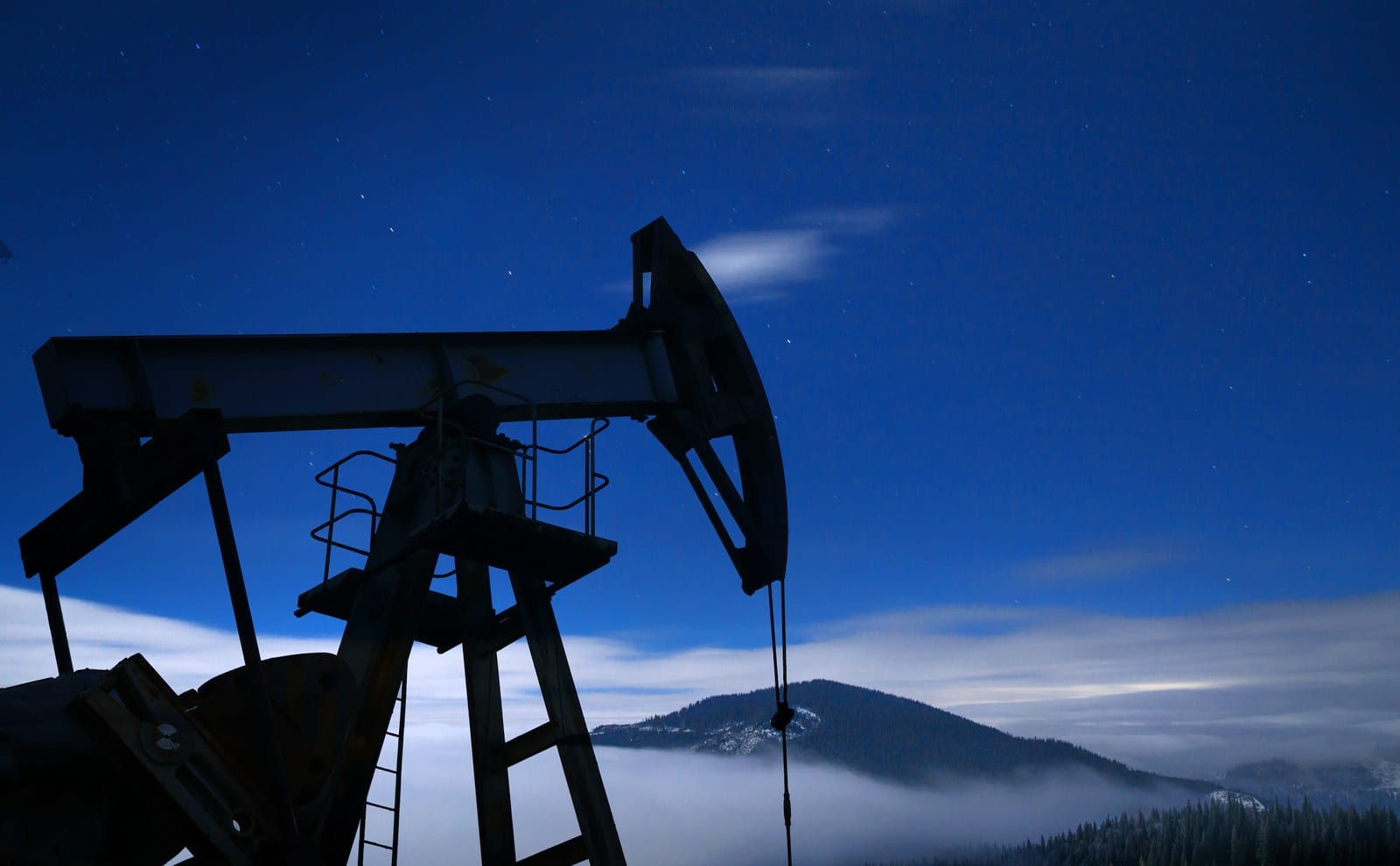Silhueta de uma bomba de extração de petróleo contra um céu azul noturno, com montanhas e névoa ao fundo