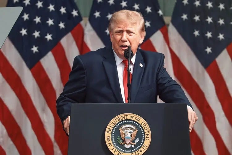 Donald Trump, de terno escuro e gravata vermelha, fala em um púlpito com o selo presidencial, diante de bandeiras dos EUA