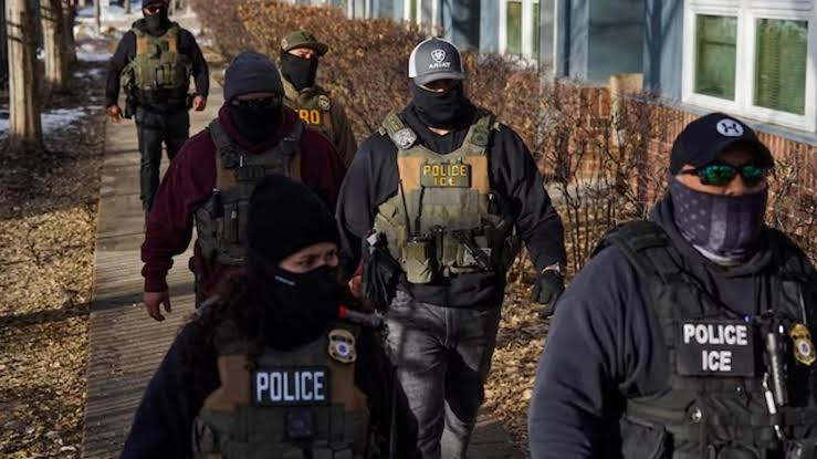 Agentes do ICE com coletes táticos com a inscrição Police ICE e toucas cobrindo o rosto caminham em fila por uma calçada durante operação de campo