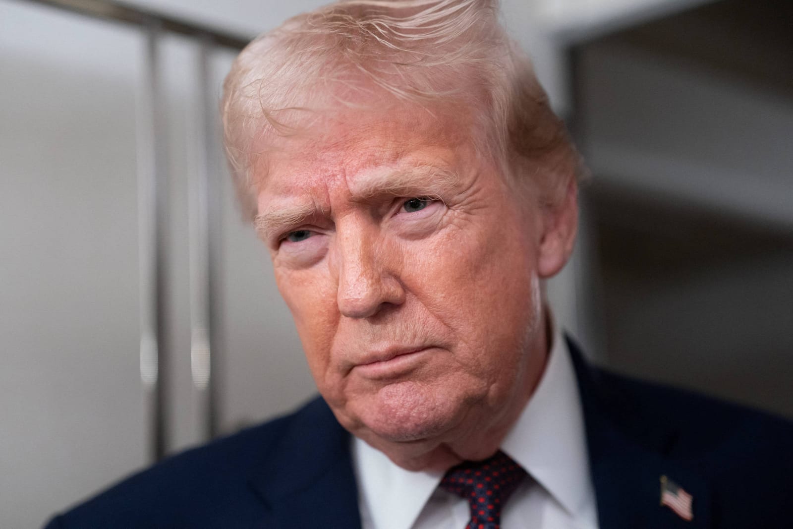 Retrato em close do presidente Donald Trump com expressão séria, de terno azul-escuro, camisa branca e gravata com pontos vermelhos