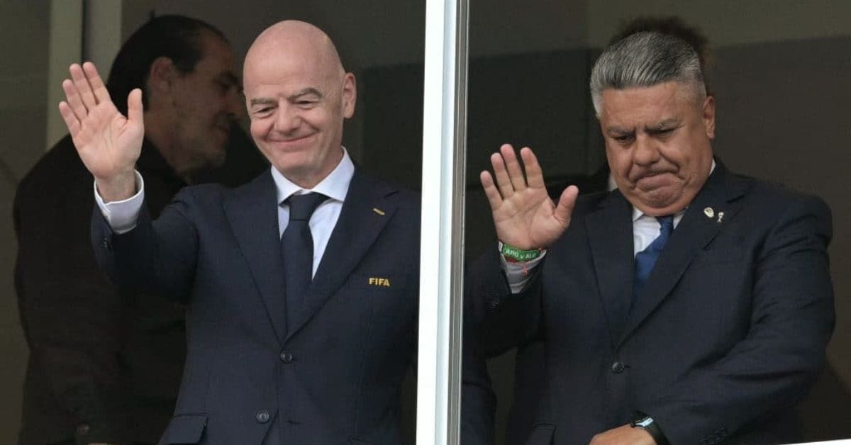 O presidente da FIFA, Gianni Infantino, e o presidente da AFA, Claudio Tapia, acenam lado a lado em um camarote de estádio, ambos de terno escuro