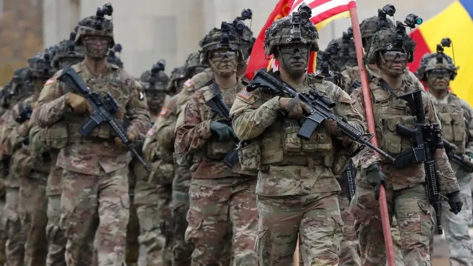 Soldados americanos em uniforme camuflado e capacete marcham em formação cerrada com fuzis, um deles carregando uma bandeira dos Estados Unidos