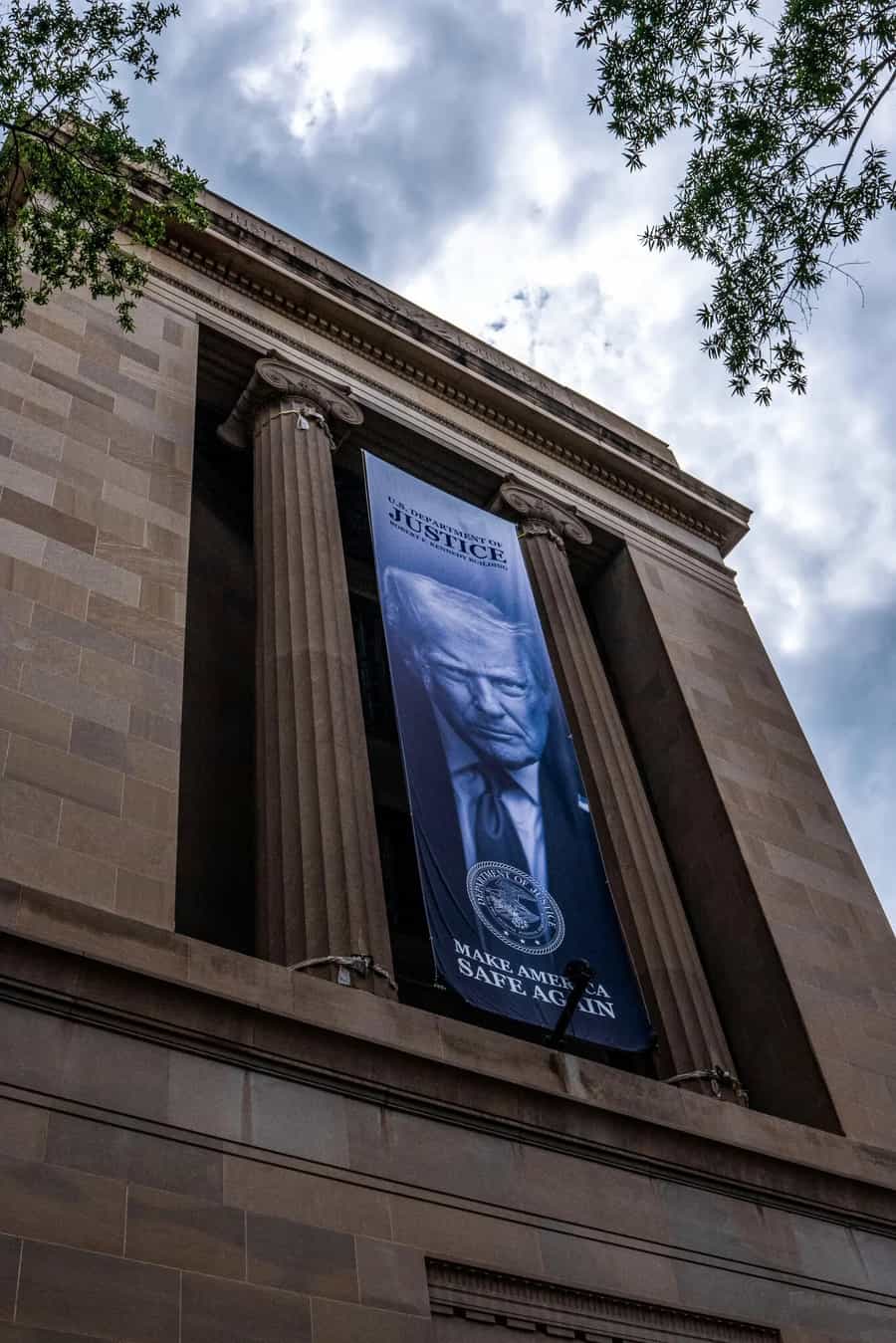 Fachada do prédio do Departamento de Justiça em Washington vista de baixo, com uma faixa vertical pendurada entre as colunas trazendo a foto do presidente Donald Trump e a frase MAKE AMERICA SAFE AGAIN