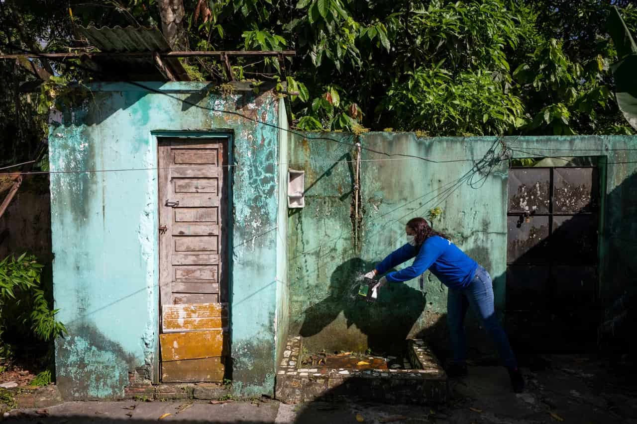Agente de saúde de blusa azul e máscara se inclina para aplicar produto em um ponto úmido junto ao muro de uma casa simples cercada pela vegetação