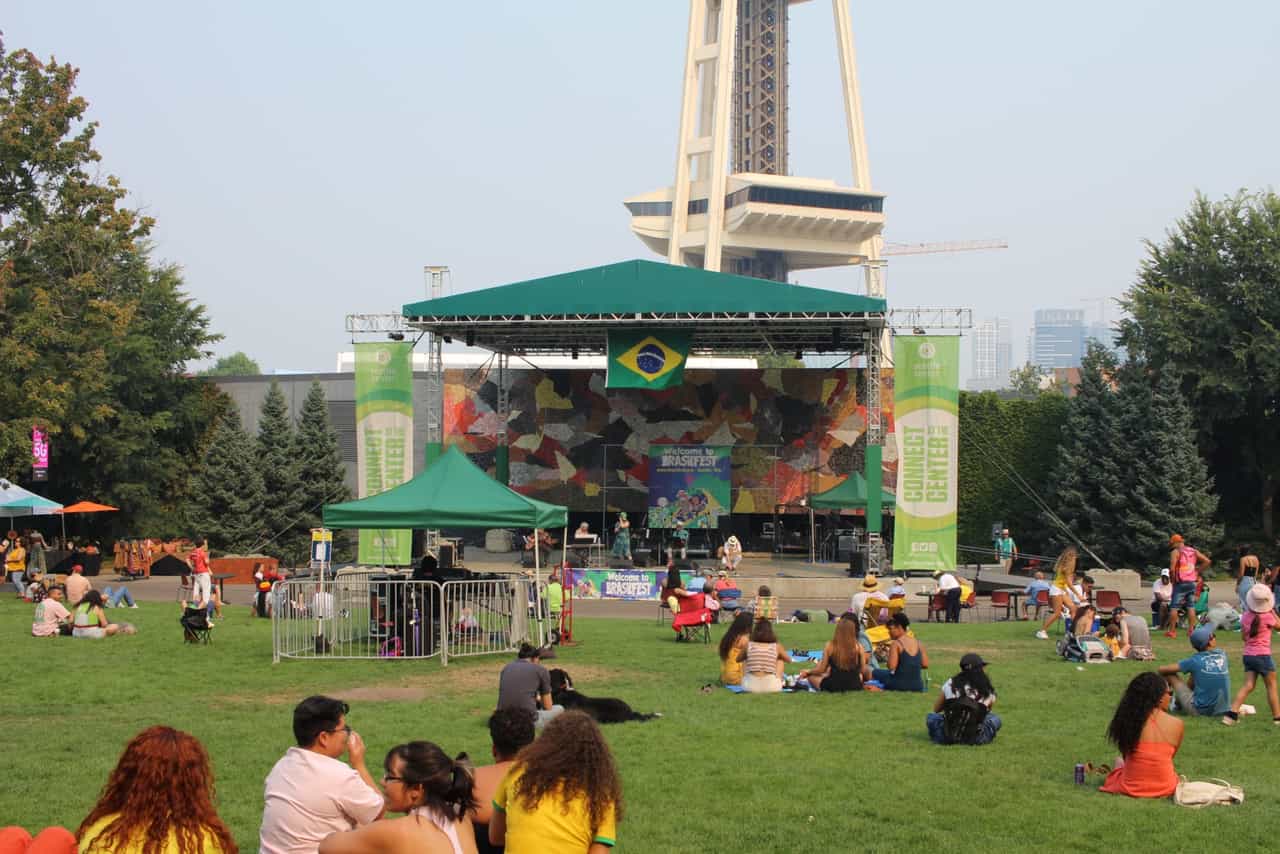 Público sentado no gramado diante do palco do BrasilFest, com bandeira do Brasil na cobertura e a Space Needle ao fundo, no Seattle Center