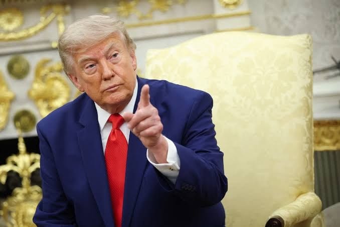 Donald Trump, de terno azul e gravata vermelha, sentado em uma poltrona dourada, aponta o dedo para a frente