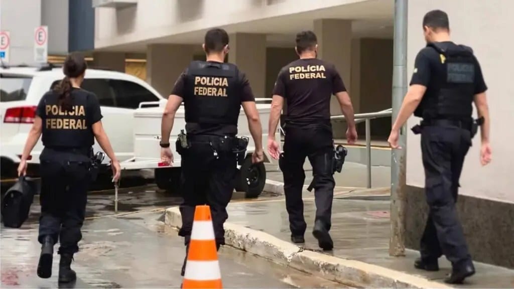 Quatro agentes da Polícia Federal, com coletes e camisas pretas, caminham em uma rua ao lado de um cone de sinalização