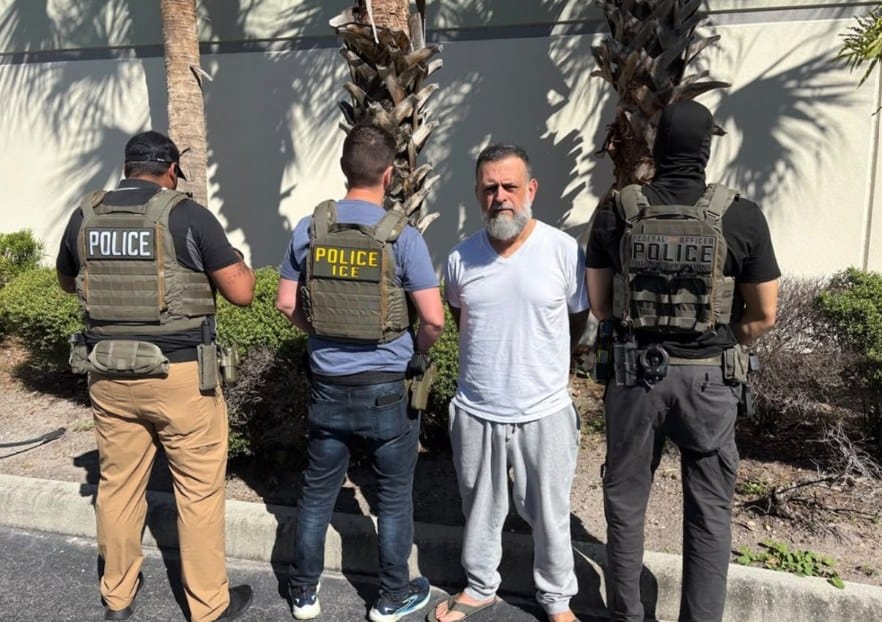 Homem de barba grisalha e camiseta branca, de costas para o muro, entre agentes de coletes escritos Police e ICE, em um estacionamento