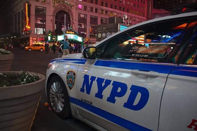 Viatura branca e azul da NYPD estacionada ao lado de uma jardineira, com as luzes de Times Square ao fundo, à noite