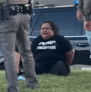 Mulher sentada na grama ao lado de agentes, um deles policial estadual do Texas, após uma detenção em via pública