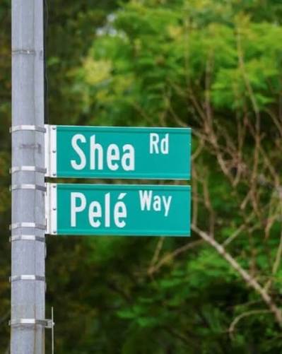 Placas de rua verdes 'Shea Rd' e 'Pelé Way' fixadas em um poste, com árvores ao fundo