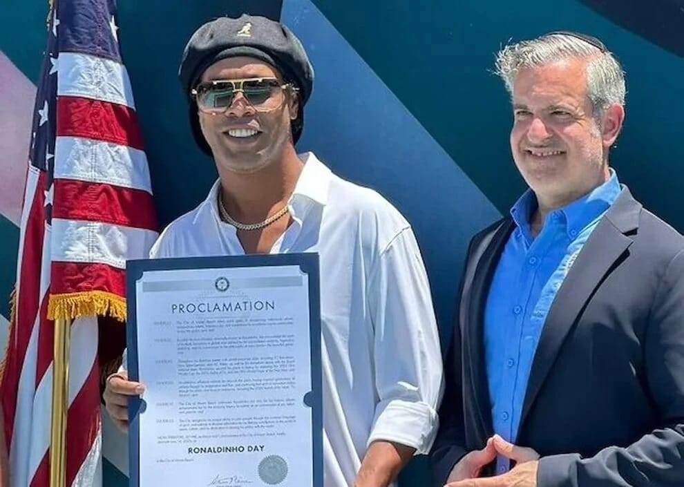 Ronaldinho, de boina e óculos escuros, segura uma proclamação 'Ronaldinho Day' ao lado do prefeito de Miami Beach, diante da bandeira dos EUA