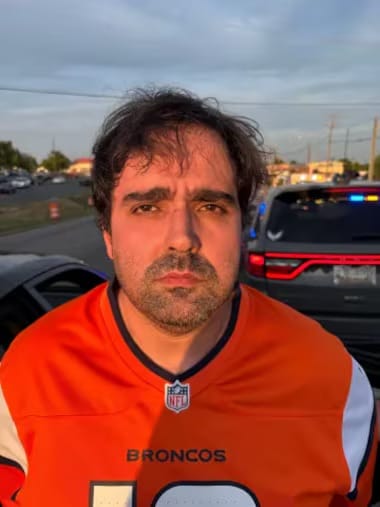 Homem de cabelos escuros com camisa laranja do Denver Broncos, com viatura policial de luzes acesas ao fundo, em estacionamento ao entardecer