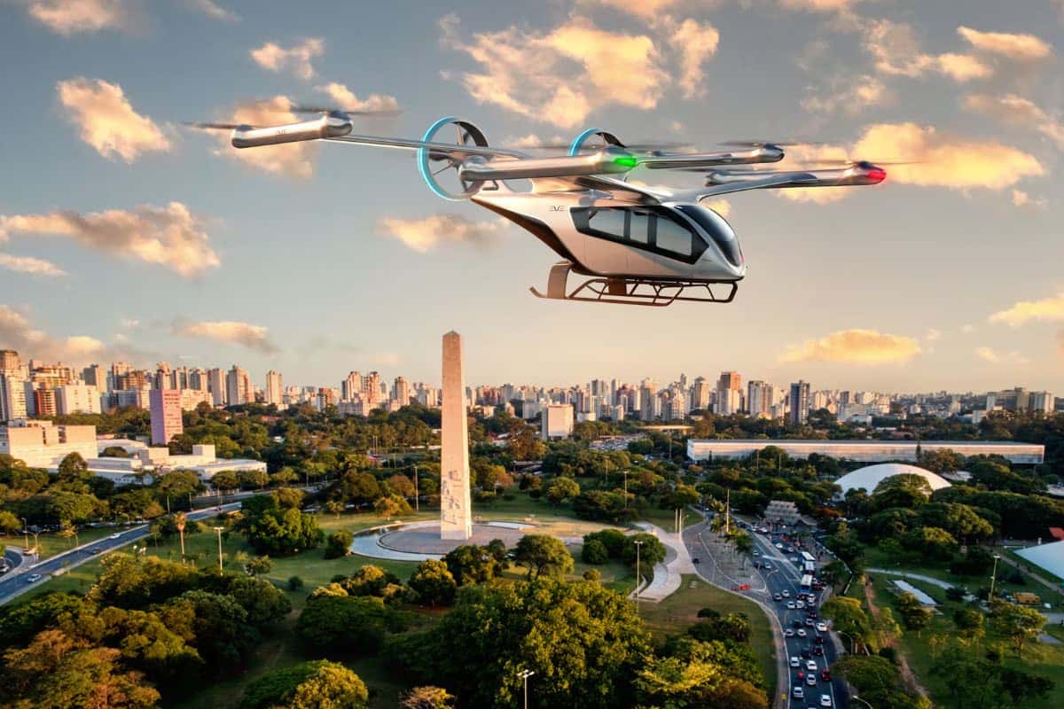 Ilustração de um eVTOL branco da Eve com oito rotores e cabine de passageiros sobrevoando um parque urbano com obelisco e a cidade ao fundo