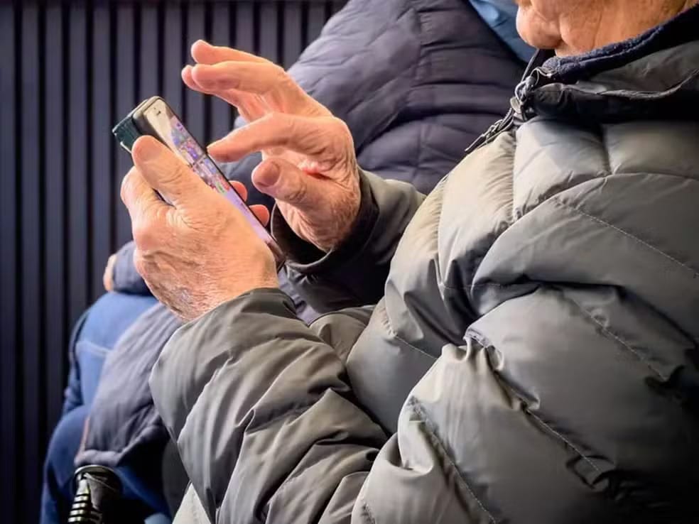 Pessoa idosa de casaco acolchoado cinza toca a tela de um smartphone com as duas mãos, vista em primeiro plano