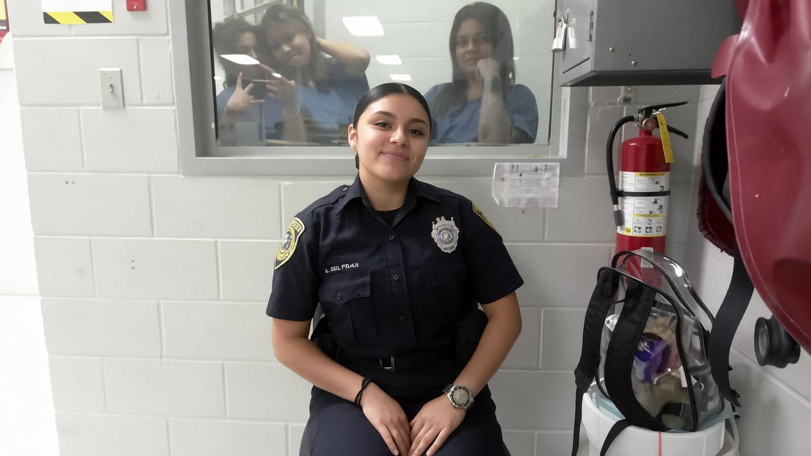 Agente de detenção de uniforme azul-escuro sorri sentada diante de um janelão de vidro, com duas detentas de uniforme azul atrás