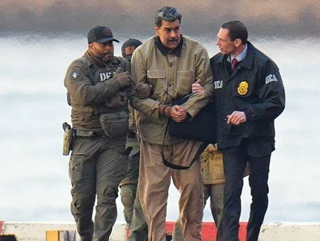Homem de casaco bege e mãos algemadas à frente é conduzido por dois agentes com coletes e jaquetas da DEA, diante do mar