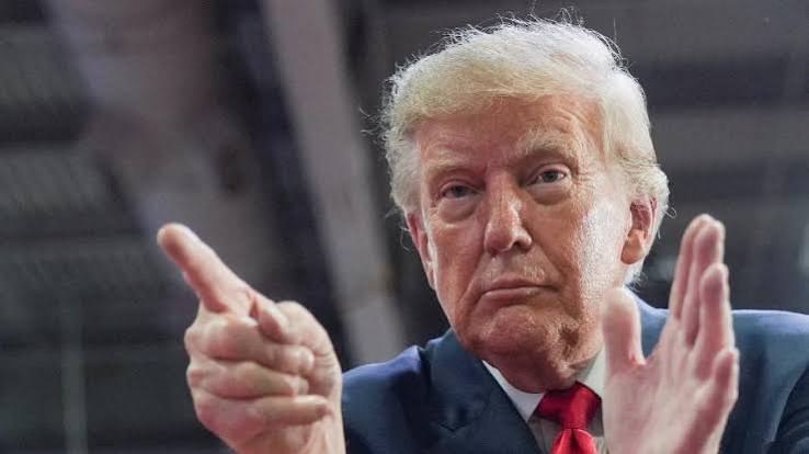Donald Trump, de terno azul e gravata vermelha, faz um gesto com as duas mãos e olha para a câmera