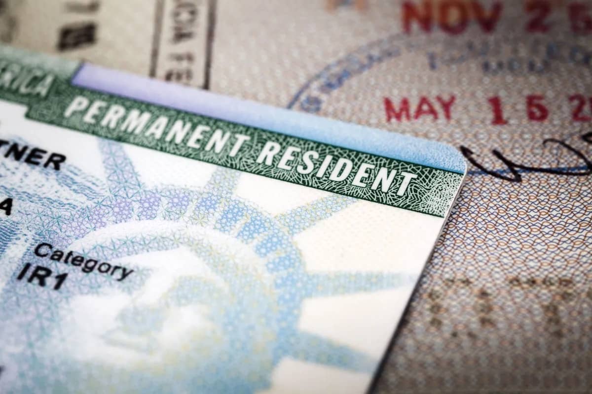 Close de um green card dos EUA com a inscrição PERMANENT RESIDENT e categoria IR1, sobre um passaporte com carimbos de imigração