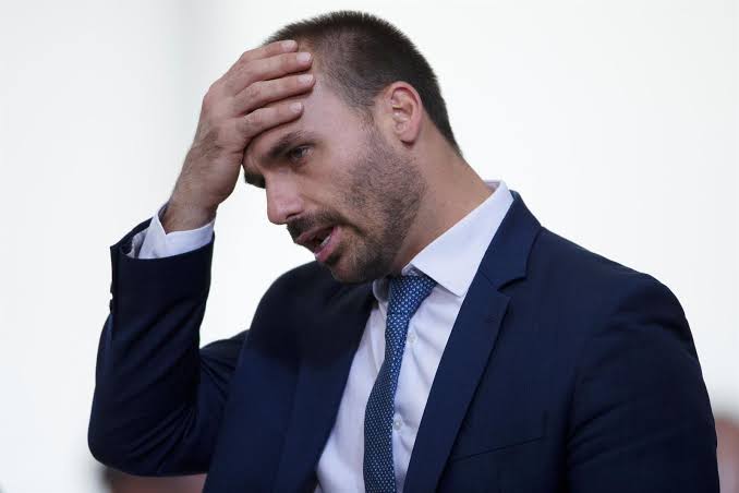 Eduardo Bolsonaro de terno azul com a mão na testa e expressão preocupada