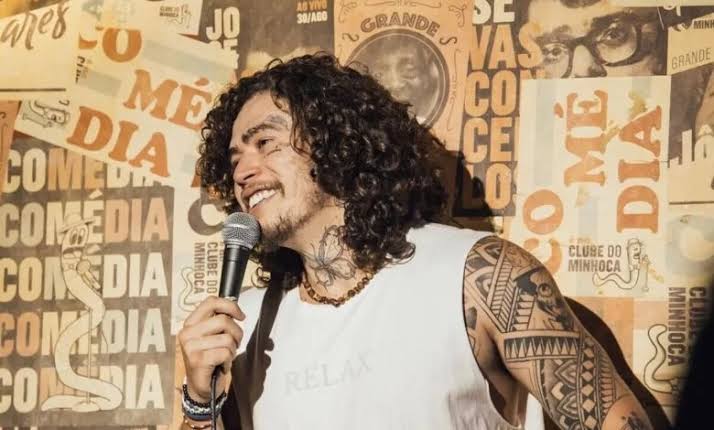 Whindersson Nunes, de cabelos cacheados e regata branca, sorri ao microfone diante de um painel com cartazes do Clube do Minhoca