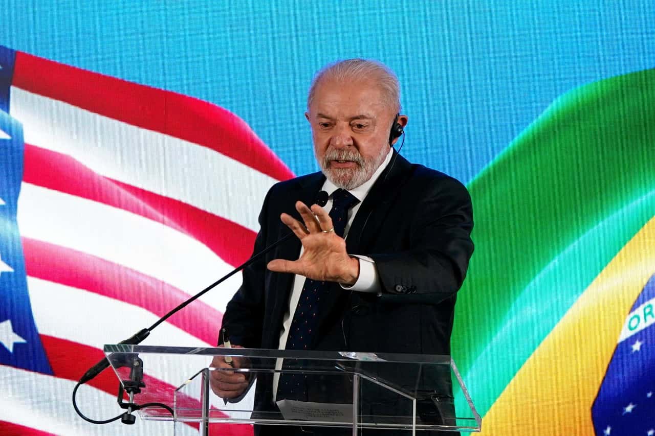 O presidente Lula, de terno escuro e fone de tradução, gesticula ao discursar em púlpito de acrílico, diante de um telão com as bandeiras dos EUA e do Brasil