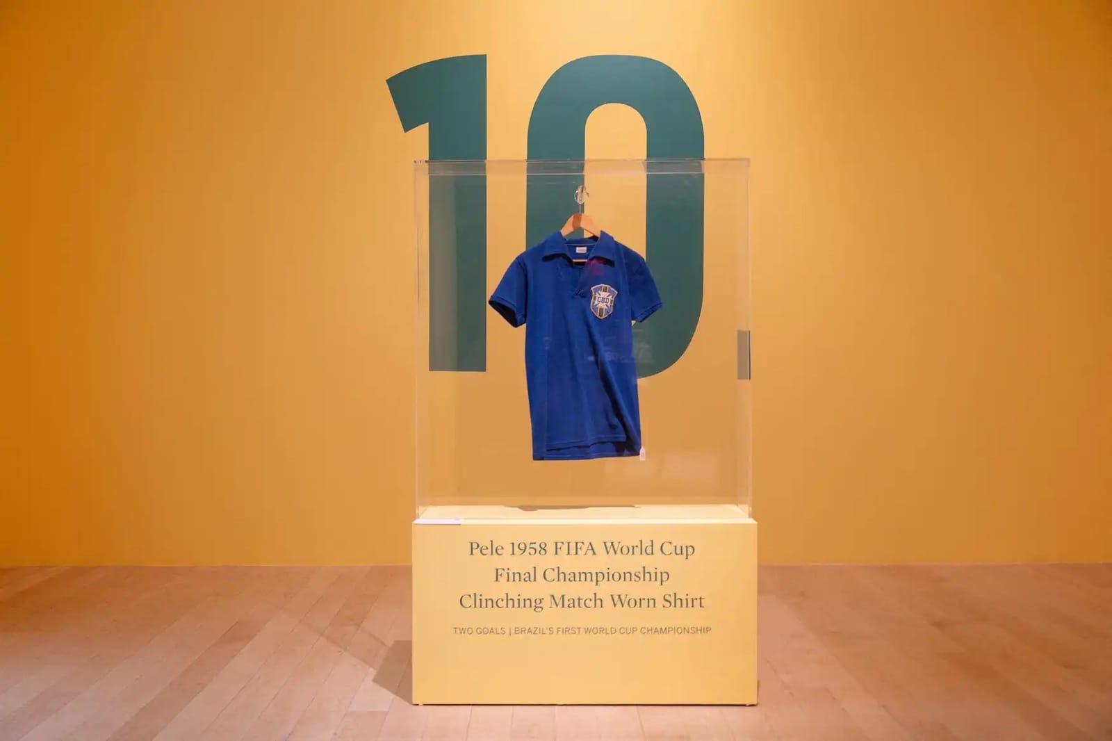 A camisa azul número 10 usada por Pelé na final da Copa de 1958, com o escudo da CBD, exposta em vitrine de acrílico diante de um grande número 10 pintado em parede amarela