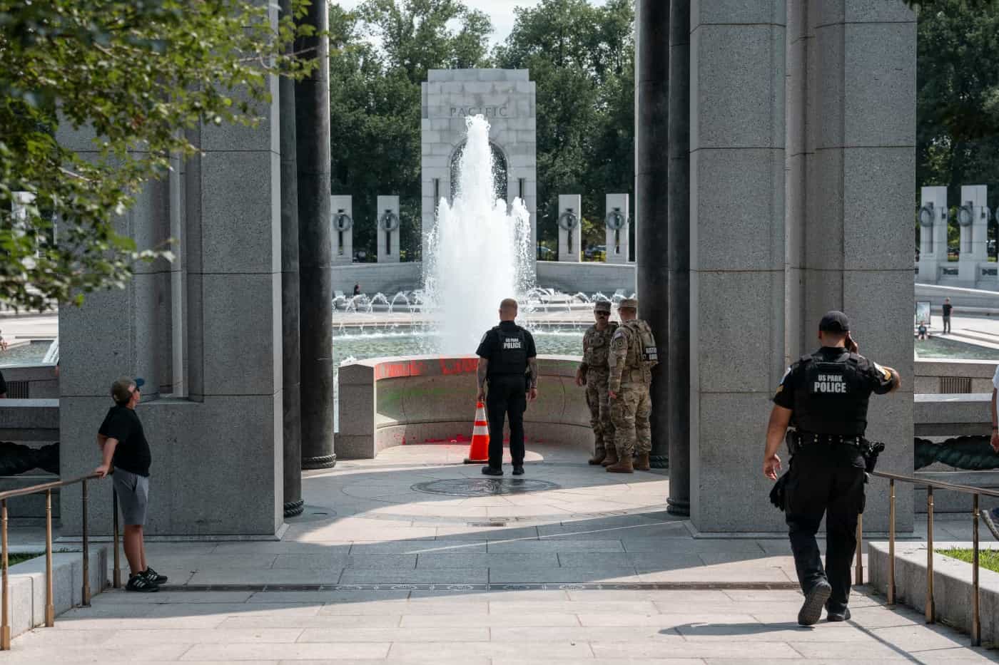 Policiais do US Park Police e dois militares da Guarda Nacional diante do muro do Memorial da Segunda Guerra Mundial marcado por tinta vermelha e verde, com a fonte do pavilhão Pacific ao fundo