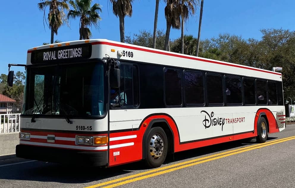 Ônibus branco e vermelho do Disney Transport, com o letreiro ROYAL GREETINGS no painel frontal, parado em via cercada de palmeiras