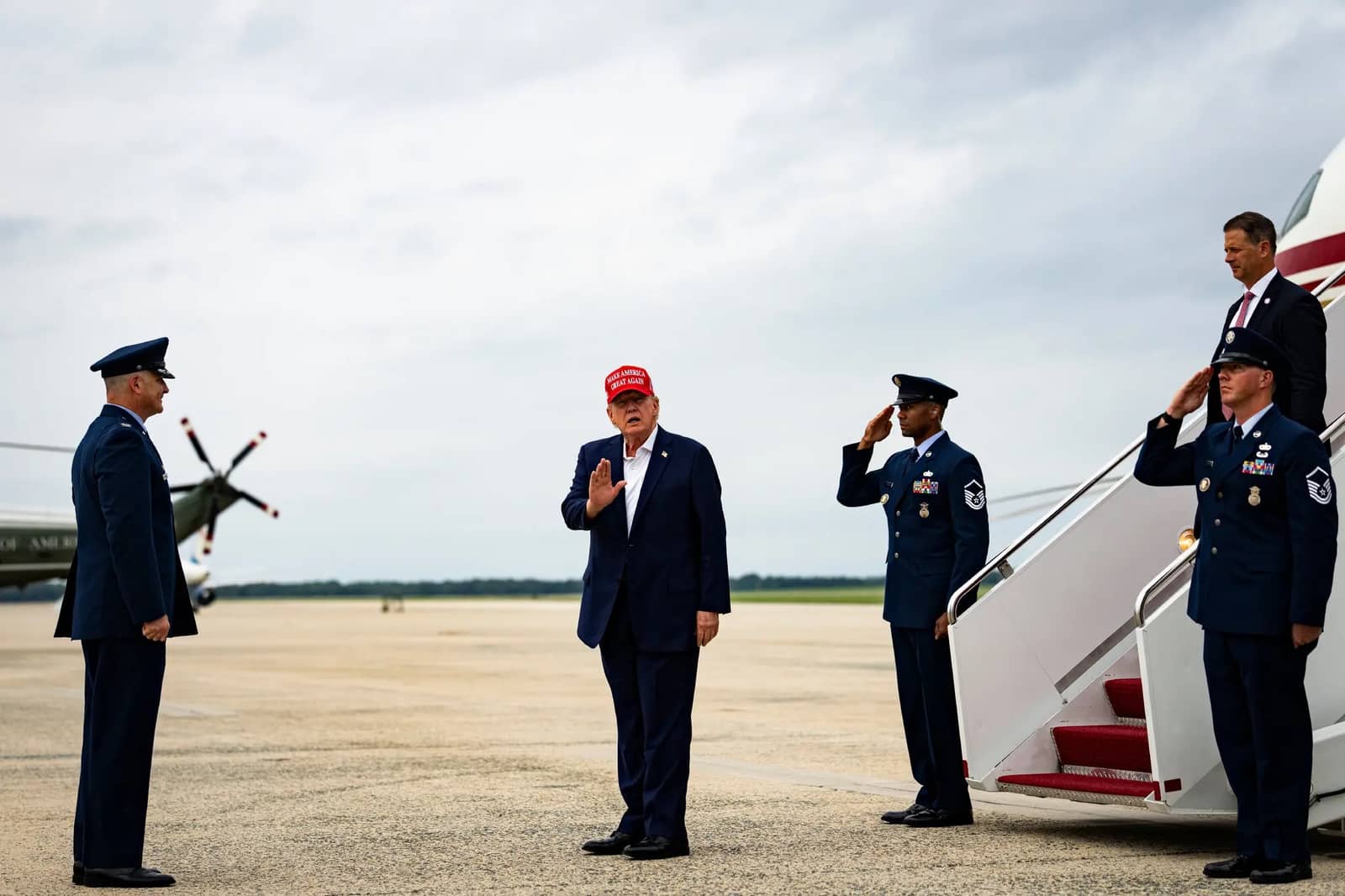 O presidente Donald Trump, de boné vermelho, ergue a mão diante de um oficial da Força Aérea enquanto dois militares o saúdam junto à escada de um avião, em pista de aeroporto