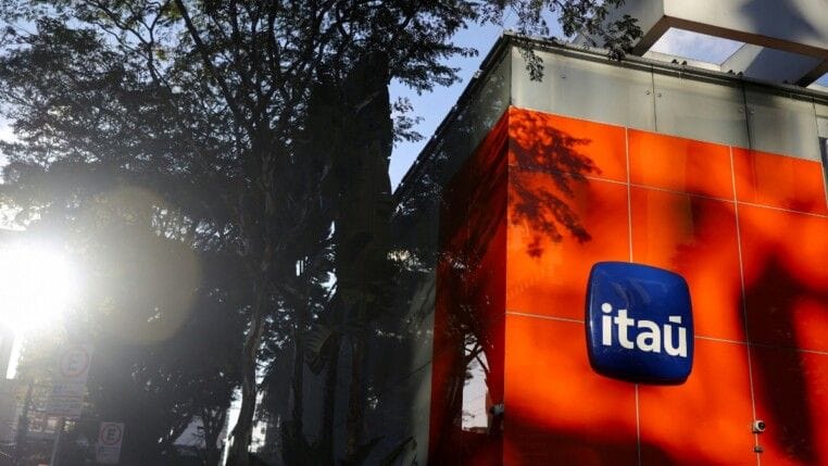 Fachada laranja de uma agência do Itaú com o logotipo em azul, vista de baixo entre as folhas de uma árvore