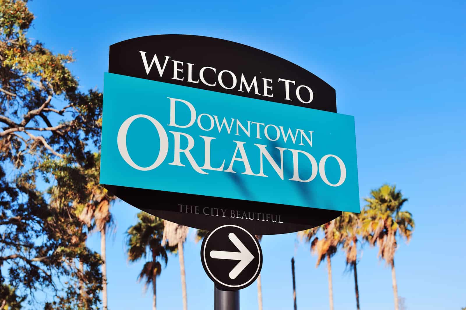 Placa de boas-vindas com os dizeres Welcome to Downtown Orlando e The City Beautiful, em azul-turquesa e preto, sob céu limpo e cercada de palmeiras