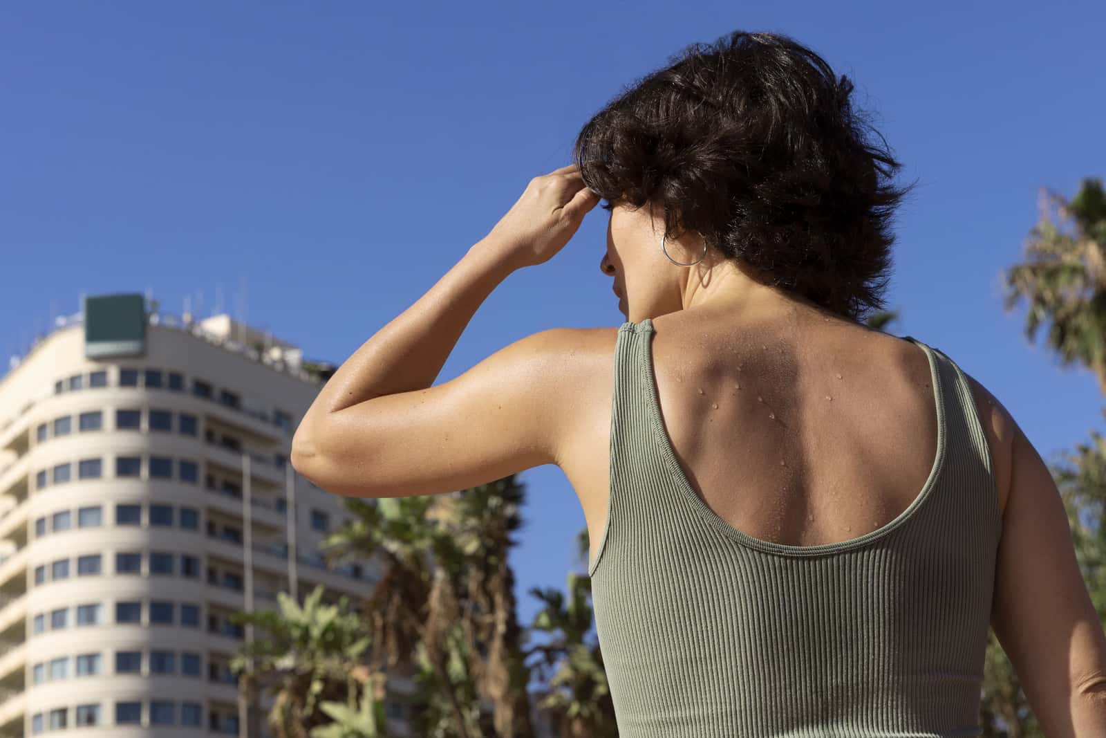Mulher de blusa verde vista de costas, com gotas de suor nos ombros, sob sol forte, diante de um edifício alto e de palmeiras