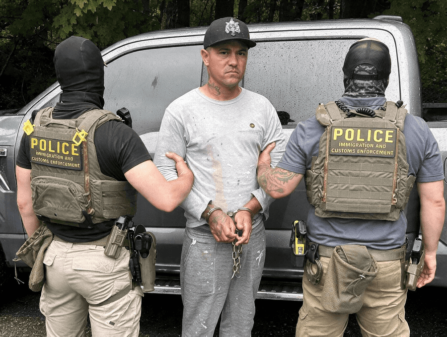 Dois agentes de coletes com a inscrição Police Immigration and Customs Enforcement conduzem um homem algemado diante de uma camionete