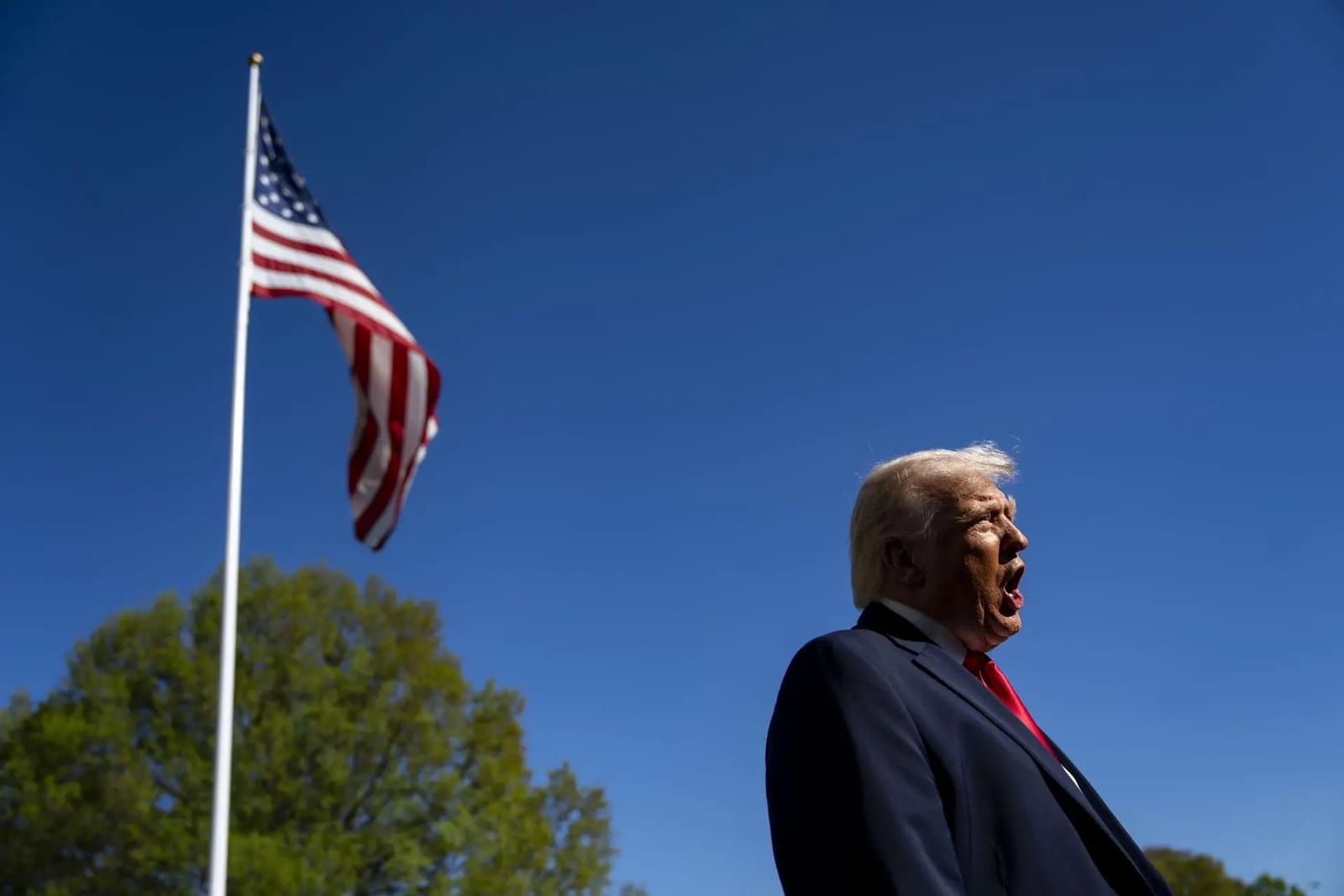 Homem de cabelos claros e terno escuro fala ao ar livre, de perfil, com a bandeira dos Estados Unidos tremulando ao fundo sob céu azul