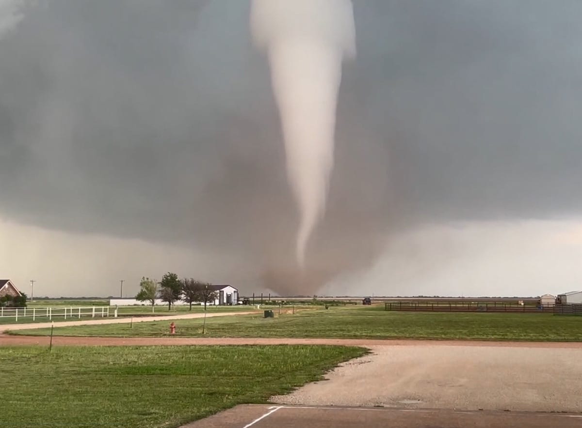 Tornado em forma de funil estreito toca o solo em área rural plana, ao lado de uma casa e cerca brancas, sob céu carregado
