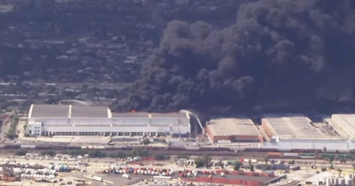Vista aérea de um grande galpão industrial tomado por uma densa coluna de fumaça preta, com bairro residencial e pátio de contêineres ao redor