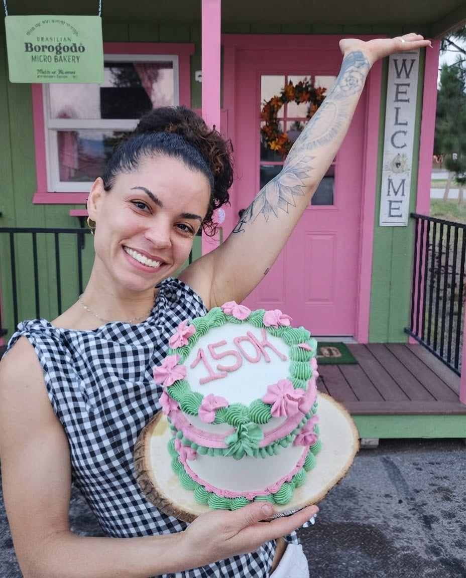 Mulher de vestido xadrez preto e branco sorri segurando um bolo decorado em verde e rosa com o número 150K, em frente a uma casinha rosa e verde com a placa Brasilian Borogodó Micro Bakery