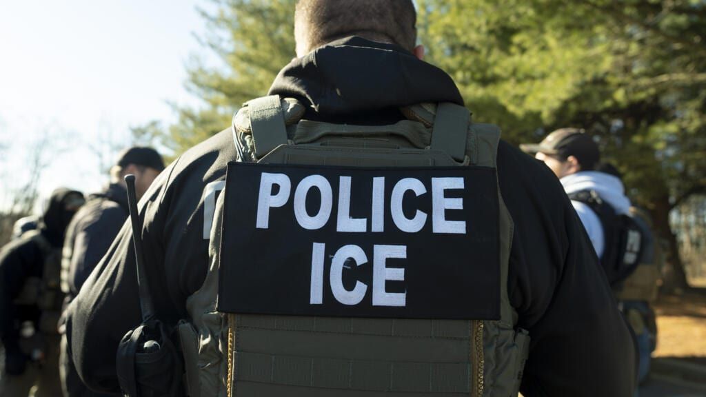 Agente visto de costas usando colete tático com a inscrição POLICE ICE em letras brancas, ao lado de outros agentes em área arborizada